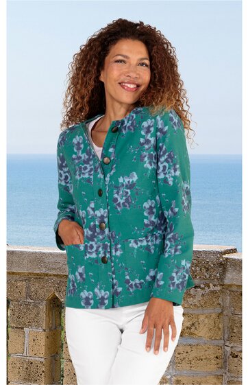 Grüne Bluse mit lila und weißen Blumenmuster, Knopfleiste vorne, lange Ärmel, taillierte Form, getragen von stehender Frau mit lockigem Haar, seitlich schräg zur Kamera gewandt