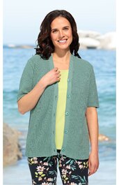 Grüne kurzärmelige Strickjacke mit V-Ausschnitt, Knöpfen vorne, getragen von lächelnder Frau, vorne offen, locker sitzend, darunter hellgelbes Oberteil, kombiniert mit dunkelblauer Hose mit großem Blumenmuster, stehende Pose vor Wasser und Felsen