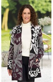 Langärmliger langer Cardigan in Grau, Schwarz und Dunkelbordeaux mit großem floralem Muster, offen, ohne Verschluss, getragen von stehender Frau mit weißen Rollkragenpullover und schwarzer Hose