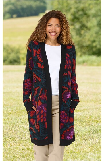 Lange Strickjacke in Dunkelblau mit buntem Blumenmuster, offene Vorderseite, lange Ärmel, zwei Eingrifftaschen, getragen von stehender Frau mit lockerem Haar, weiße Bluse darunter, beige Hose