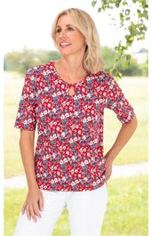 Rotes Damenoberteil mit weißem und blauem Blumenmuster, rundem Ausschnitt mit kleinem Schlitz und Knopf, kurze Ärmel, gerade geschnitten, getragen von stehender Frau mit linker Hand in Hüfte, heller Hintergrund im Freien