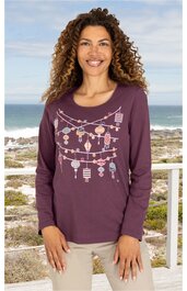 Langärmeliges T-Shirt in dunklem Violett mit buntem Laternen-Motiv auf der Vorderseite, rundem Halsausschnitt, gerade geschnitten, getragen von stehender Frau vor Küstenlandschaft
