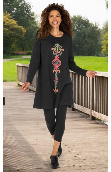 Schwarzes, langärmliges Tunikakleid mit farbigem, vertikalem Muster vorne, rundem Ausschnitt, knieumspielend, getragen von lächelnder Frau mit lockigen Haaren, kombiniert mit schwarzer Leggings und schwarzen Schuhen, stehend an Holzbrüstung im Freien