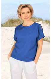 Blaues Damenoberteil mit kurzen Ärmeln, Rundhalsausschnitt, schulterfreie Aussparung rechts, lockere Passform, getragen von stehender Frau am Strand, weißer locker sitzender Hose