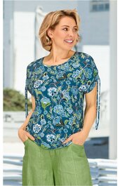 T-Shirt, blau mit floralen Mustern in Weiß, Hellblau und Grün, rundem Ausschnitt, kurze Ärmel mit Bindebändern, getragen von lächelnder stehender Frau, legerer Schnitt