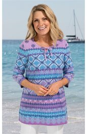 Langärmliges Tunika-Oberteil in Helllila, Hellblau und Türkis mit geometrischem Muster, Rundhalsausschnitt mit kleinen Bändern zum Binden, gewellter Saum, getragen von lächelnder Frau am Meer, locker geschnitten