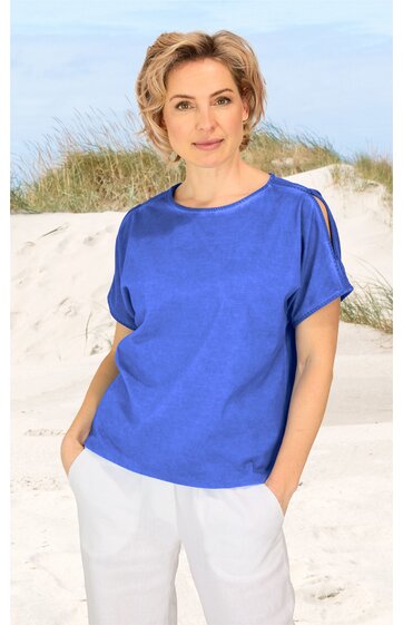 Blaues Damenoberteil mit kurzen Ärmeln, Rundhalsausschnitt, schulterfreie Aussparung rechts, lockere Passform, getragen von stehender Frau am Strand, weißer locker sitzender Hose