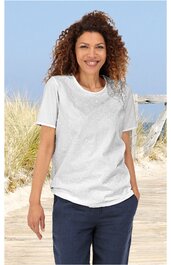 Weißes T-Shirt mit floralem Muster auf der linken Schulter, rundem Ausschnitt, kurze Ärmel, locker sitzend, getragen von stehender Frau vor Strandhintergrund, dunkelblaue Hose