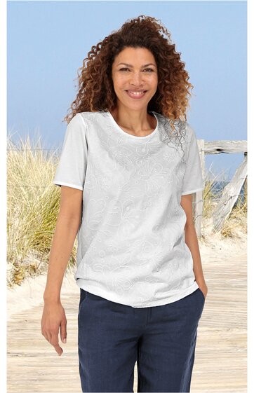 Weißes T-Shirt mit floralem Muster auf der linken Schulter, rundem Ausschnitt, kurze Ärmel, locker sitzend, getragen von stehender Frau vor Strandhintergrund, dunkelblaue Hose