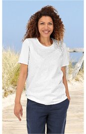 Weißes T-Shirt mit floralem Muster auf der linken Schulter, rundem Ausschnitt, kurze Ärmel, locker sitzend, getragen von stehender Frau vor Strandhintergrund, dunkelblaue Hose