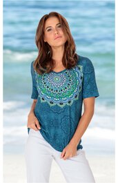Kurzärmliges T-Shirt in petrol mit großflächigem, rundem Mandala-Muster in Hellblau, Grün und Weiß auf der Vorderseite, Rundhalsausschnitt, leicht taillierte Form, getragen von stehender Frau vor Strandkulisse