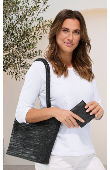 Schwarze Handtasche mit geflochtenem Muster, rechteckig, lange Henkel, gehalten von stehender Frau in weißem Langarmshirt, kombiniert mit schwarzem Portemonnaie in rechter Hand
