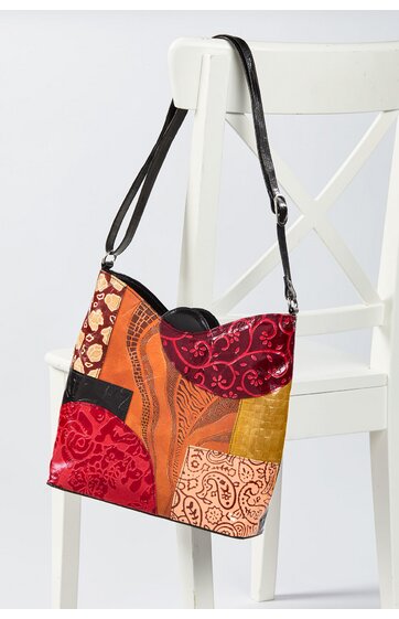 Umhängetasche in Rot-, Orange- und Beigetönen mit verschiedenen abstrakten Mustern, rechteckige Form, verstellbarer schwarzer Schulterriemen, sitzend an Rückenlehne eines weißen Stuhls