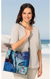 Beuteltasche, blau mit Paisley- und Patchworkmuster in verschiedenen Blautönen und Lila, rechteckige Form, breite schwarze Schulterriemen, getragen von lächelnder Frau in beigem kurzärmligem Oberteil und heller Hose am Strand