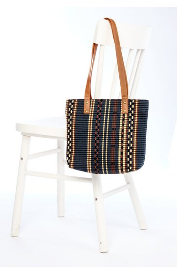 Schwarze Umhängetasche mit buntem gestreiftem Muster in Blau, Weiß, Rot und Beige, rechteckige Form, braune Schulterriemen, lose überlehnten weißen Holzstuhl hängend