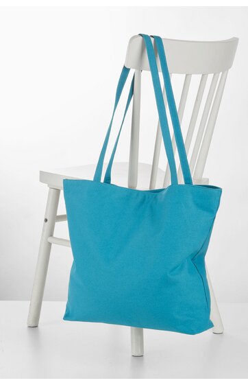 Türkisfarbene Einkaufstasche ohne Muster, rechteckige Form, lange schmale Henkel, aufgehängt an weißem Holzstuhl mit Lehne und waagerechten Stäben