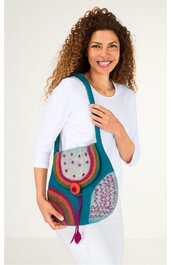 Schultertasche in Blau und Rot mit buntem Muster aus Kreisen und Linien, runde Form, roter Knopfverschluss, langer Schulterriemen, getragen von lächelnder Frau in weißem Langarmshirt und weißer Hose, Tasche aus Stoff