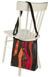 Handtasche mit aufrechten, schmalen Streifen in Rot, Orange und Schwarz, geometrisches Muster, längenverstellbarer schwarzer Schulterriemen, trapezförmige Form, hängend an Rückenlehne eines weißen Holzstuhls
