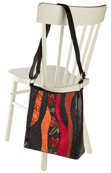 Handtasche mit aufrechten, schmalen Streifen in Rot, Orange und Schwarz, geometrisches Muster, längenverstellbarer schwarzer Schulterriemen, trapezförmige Form, hängend an Rückenlehne eines weißen Holzstuhls