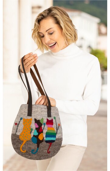 Graue Umhängetasche mit buntem Katzenmotiv in orange, türkis, pink und blau, zwei dunkelbraune Tragegriffe, an runder Form, getragen von lächelnder Frau im weißen Rollkragenpullover