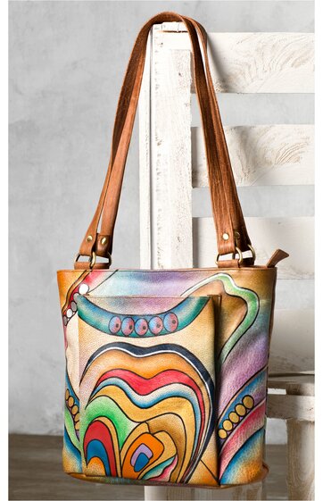 Bunte Umhängetasche mit abstraktem Muster in Grün, Gelb, Rot und Blau, rechteckige Form, zwei lange braune Henkel, sichtbare Nähte, Tasche steht auf weißem Holzstuhl vor grauer Wand