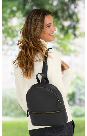 Schwarzer Rucksack mit zwei Reißverschlüssen, glatte Oberfläche, schlichter Look, Rückenansicht, getragen von lachender Frau mit langem braunem Haar und weißem Oberteil, draußen in grüner Umgebung