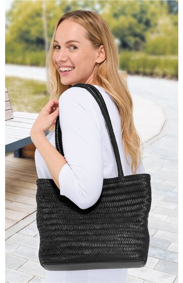 Schwarze, geflochtene Handtasche mit langen Henkeln, rechteckige Form, getragen von Frau in weißem Oberteil, Outdoor-Umgebung mit gepflastertem Boden und Tisch