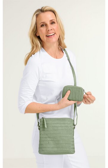 Olivgrüne Umhängetasche mit Flechtmuster, rechteckig, mit Reißverschluss oben, längenverstellbarer Schultergurt, dazu passende kleine Geldbörse, getragen von lächelnder Frau in weißem Oberteil und weißer Hose