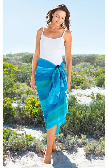 Weißes Top, ärmellos, Schulterträger, knöchellanger blauer Pareo mit Streifenmuster, gewickelt und vorne gebunden, getragen von lächelnder Frau, stehend im Freien auf sandigem Boden mit grünem Gestrüpp