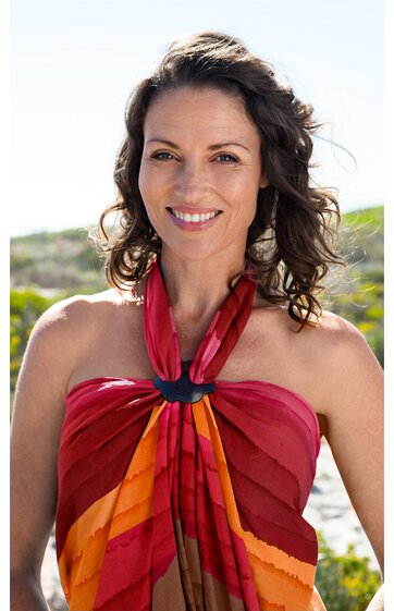 Frau mit mittellangen lockigen braunen Haaren, trägt mehrfarbiges Strandkleid in Rot-, Orange- und Brauntönen mit schwarzem Ring am Hals, ärmellos, am Strand mit Sand und Meer im Hintergrund