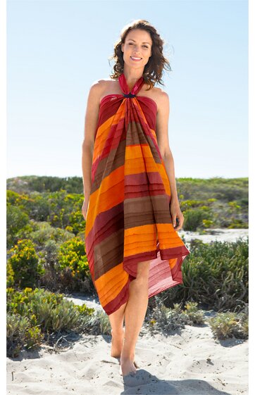 Strandkleid in Orange- und Brauntönen, gestreift, asymmetrischer Schnitt, gebunden am Hals, ärmellos, knielang, getragen von stehender Frau im Sand mit grünen Büschen im Hintergrund