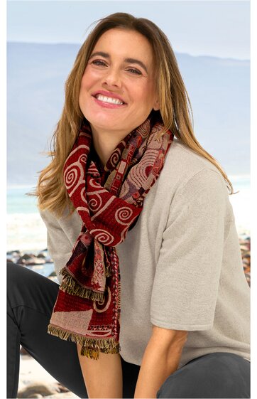 Schal mit rot-beigem Ethno-Muster, Fransen an den Enden, getragen von lächelnder Frau mit beige-grauem Kurzarmshirt, sitzend, helle Outdoor-Umgebung