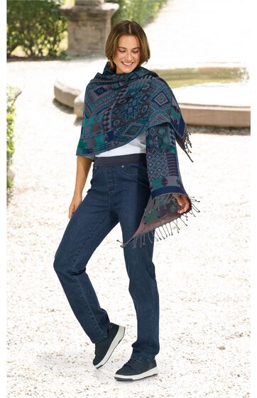 Blau-grüner Schal mit geometrischem Muster und Fransen, um die Schultern einer stehenden Frau gelegt, kombiniert mit dunkelblauen Jeans und blauen Sneakers