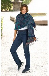 Blau-grüner Schal mit geometrischem Muster und Fransen, um die Schultern einer stehenden Frau gelegt, kombiniert mit dunkelblauen Jeans und blauen Sneakers