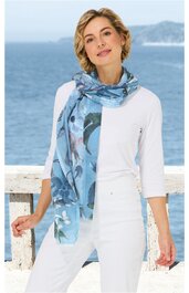 Langer, blau gemusterter Schal mit abstracten Motiven, getragen locker um den Hals, dazu weiße, dreiviertelärmlige Bluse, weiße Hose, hellblaue Sneaker, stehende Person mit locker gesenktem Kopf und leichtem Lächeln