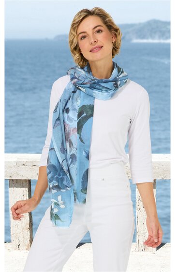 Langer, blau gemusterter Schal mit abstracten Motiven, getragen locker um den Hals, dazu weiße, dreiviertelärmlige Bluse, weiße Hose, hellblaue Sneaker, stehende Person mit locker gesenktem Kopf und leichtem Lächeln