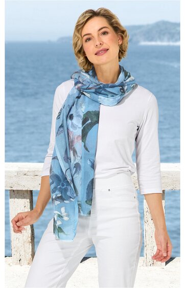 Langer, blau gemusterter Schal mit abstracten Motiven, getragen locker um den Hals, dazu weiße, dreiviertelärmlige Bluse, weiße Hose, hellblaue Sneaker, stehende Person mit locker gesenktem Kopf und leichtem Lächeln