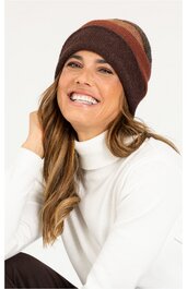 Braune Strickmütze mit dunkleren und helleren braunen Streifen, eng anliegend, getragen von lächelnder Frau mit langen gewellten Haaren, weißer Rollkragenpullover, sitzend, linke Hand am Kopf