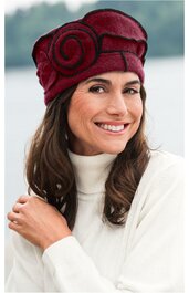 Bordeauxroter Damenhut mit großer, geschwungener Spiraldekoration, eng anliegend, getragen von lächelnder Frau mit langen braunen Haaren und weißem Rollkragenpullover