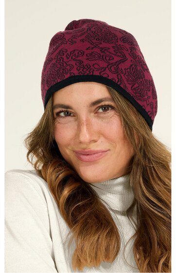 Dunkelrote Beanie-Mütze mit schwarzem Blumenmuster, schwarzer Umschlagkante, getragen von lächelnder Frau mit langen, welligen, hellbraunen Haaren, weißer Rollkragenpullover