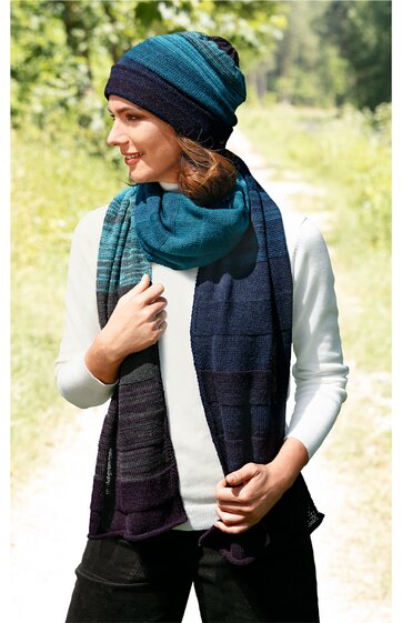 Damenmütze und Schalset in Blau- und Grüntönen, gestreiftes Design, locker sitzende Beanie-Mütze, langer, breiter Schal, getragen von stehender Frau mit weißem Pullover und schwarzer Hose