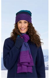 Damenmütze und Schal in lila und dunkelblau, beidseitig getragen, kombiniert mit dunkelblauem Mantel, gestrickt, Schal mit Blockstreifenmuster, getragen von lächelnder Frau mit lockigen Haaren, stehend vor winterlicher Berglandschaft