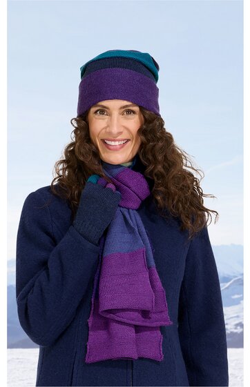 Damenmütze und Schal in lila und dunkelblau, beidseitig getragen, kombiniert mit dunkelblauem Mantel, gestrickt, Schal mit Blockstreifenmuster, getragen von lächelnder Frau mit lockigen Haaren, stehend vor winterlicher Berglandschaft