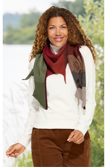 Weißer Pullover mit Rundhalsausschnitt, getragen von stehender Frau, braune Cordhose, mehrfarbiger Schal mit Streifenmustern in Rot-, Braun- und Grautönen, lange lockige Haare