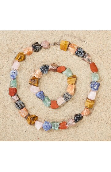 Halskette und Armband aus mehrfarbigen, unregelmäßig geformten Steinen, Braun-, Rot-, Blau-, Grün- und Schwarztöne, auf hellem Sand ausgelegt
