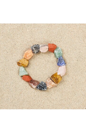 Armband aus unregelmäßigen farbigen Steinen in Braun-, Rot-, Blau-, Gelb- und Grüntönen, rund geformt, auf hellem Sand liegend