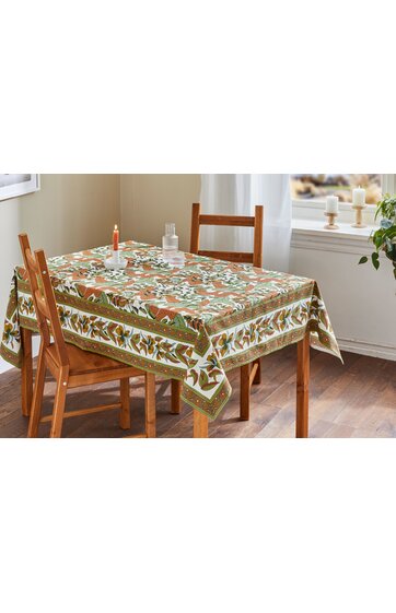 Rechteckiger Esstisch mit Tischdecke in Beige, Braun und Grün, florales Muster mit Blättern, Holzstuhl mit gerader Rückenlehne, zwei Stühle an Tisch, Kerzen und Glas auf Tisch, Fenster mit Licht im Hintergrund