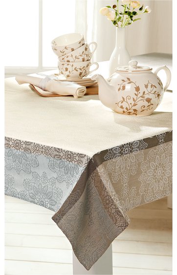 Tischdecke in Beige und Grau mit floralem Muster, rechteckig, auf Tisch ausgelegt, Kombination aus hellen und dunklen Flächen, mit Blumenornamenten, im Hintergrund weiße Teekanne und gestapelte Teetassen auf Teller, weiße Vase mit gelbweißen Blumen