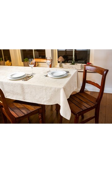 Esszimmertisch mit hellbeigem Tischläufer, gedeckt mit weißen Tellern, Gläsern, Silberbesteck auf Bastuntersetzern, umgeben von dunkelbraunen Holzstühlen mit Rückenlehne