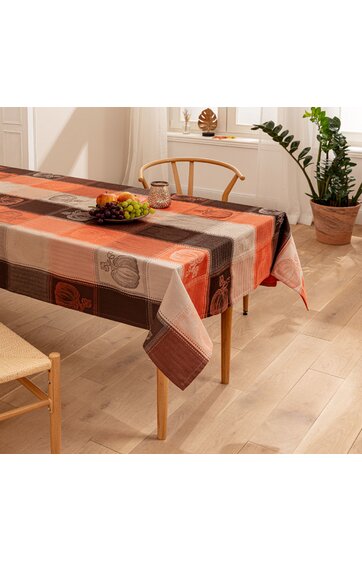 Rechteckiger Tisch mit orange-braun-beiger Tischdecke mit Kürbismotiven gemustert, Holzstuhl mit Holzlehne, Obstteller mit Trauben und Äpfeln auf dem Tisch, heller Holzboden, weiße Wände, grüner Zimmerpflanzen in Tontopf